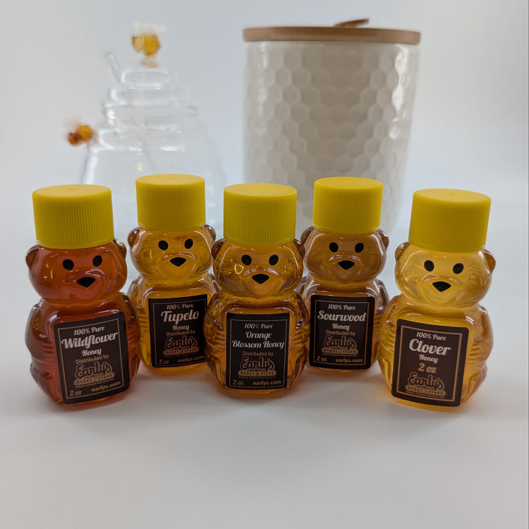 Five small bear-shaped honey bottles with yellow caps and black labels are arranged in a row on a white surface. 