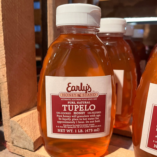 A clear plastic squeeze bottle with a bright yellow label, filled with golden-colored honey. The label features the Early's Honey Stand logo and text reading "100% Pure Tupelo Honey." The bottle is resting on a wooden table against a blurred background of honeycomb cells and a jar of honey.