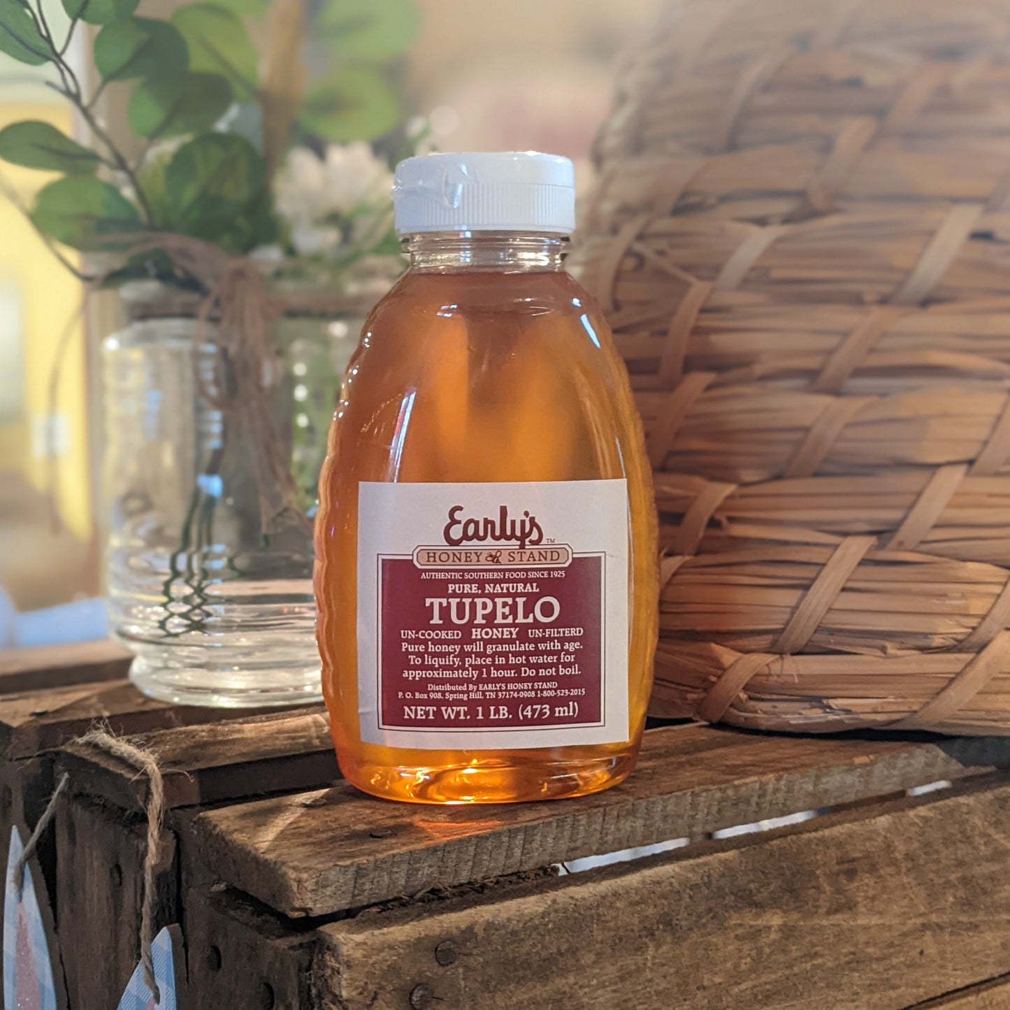 A clear plastic squeeze bottle with a bright yellow label, filled with golden-colored honey. The label features the Early's Honey Stand logo and text reading "100% Pure Tupelo Honey." The bottle is resting on a wooden table against a blurred background of honeycomb cells and a jar of honey.