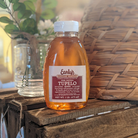 A clear plastic squeeze bottle with a bright yellow label, filled with golden-colored honey. The label features the Early's Honey Stand logo and text reading "100% Pure Tupelo Honey." The bottle is resting on a wooden table against a blurred background of honeycomb cells and a jar of honey.